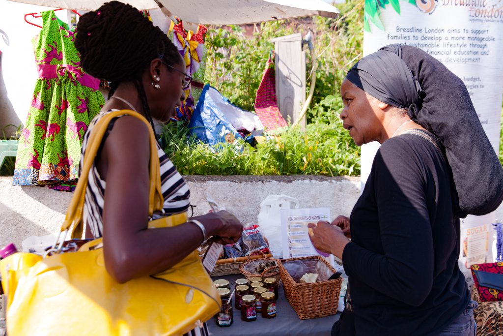 Two black women stand in front of a market stall for the social enterprise Breadline London - which aims to support families to raise themselves out of the cycle of poverty through financial education, training, practical workshops, and business opportunities.